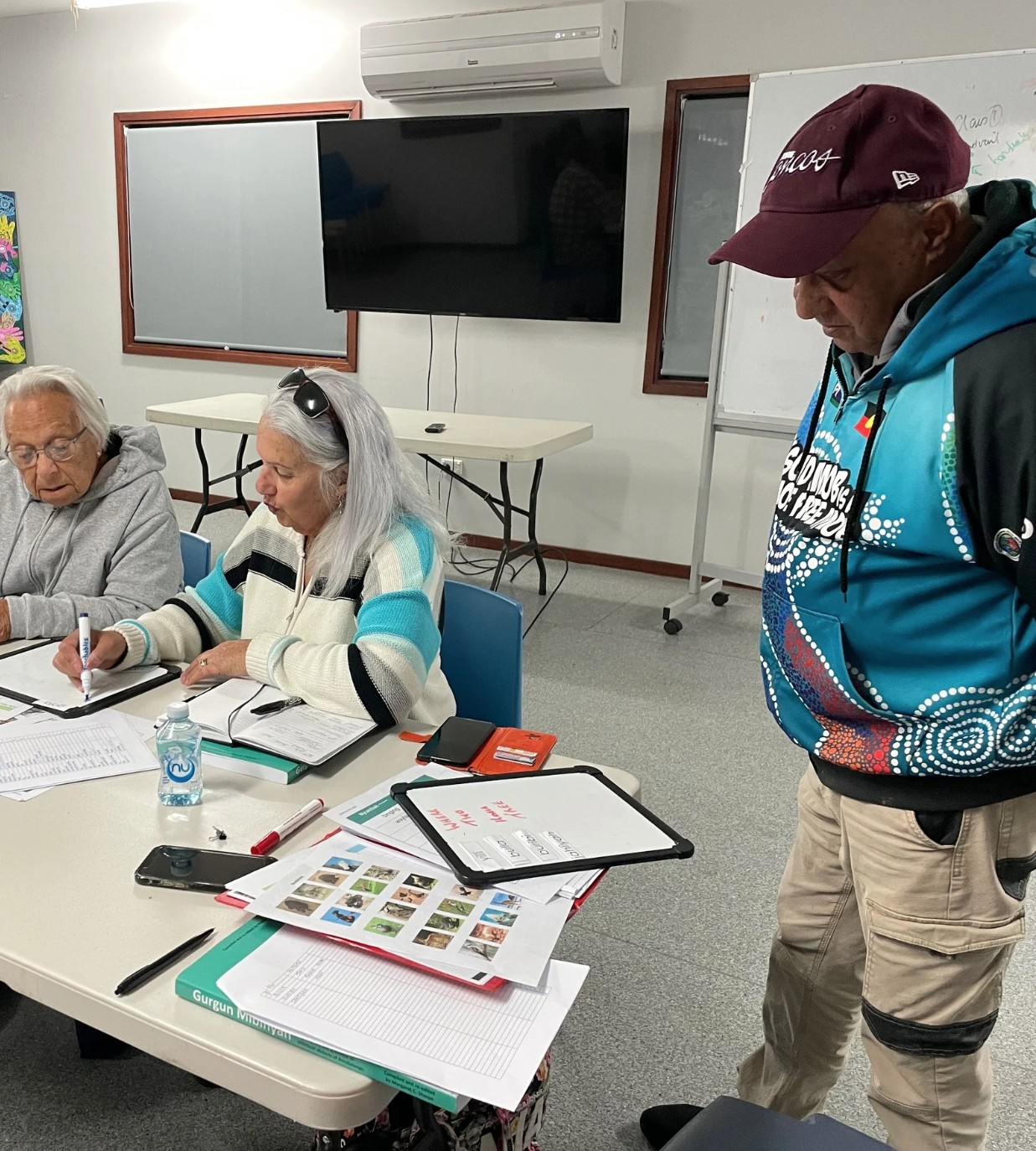 Bundjalung Elders and community members sit around a table during a language research session, reviewing animal reference photos, notes, and educational resources. One Elder writes in a notebook while others stand nearby, contributing to the collaborative process of reconnecting and documenting Bundjalung cultural knowledge.